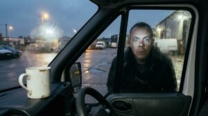A wet depot yard before dawn as seen from inside a van cab: a chipped white mug sits on the dashboard, and just outside the rain-spotted window a man in a dark hood presses his face to the glass, eyes wide, peering in. Sodium lights glare on the wet tarmac beyond. No one else is in the frame.